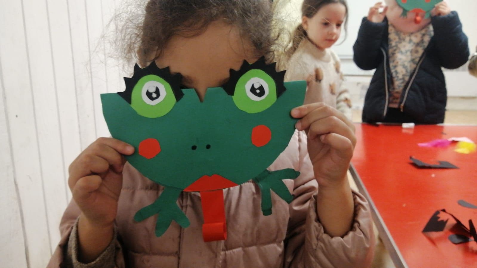 A child hides behind a paper frog mask.