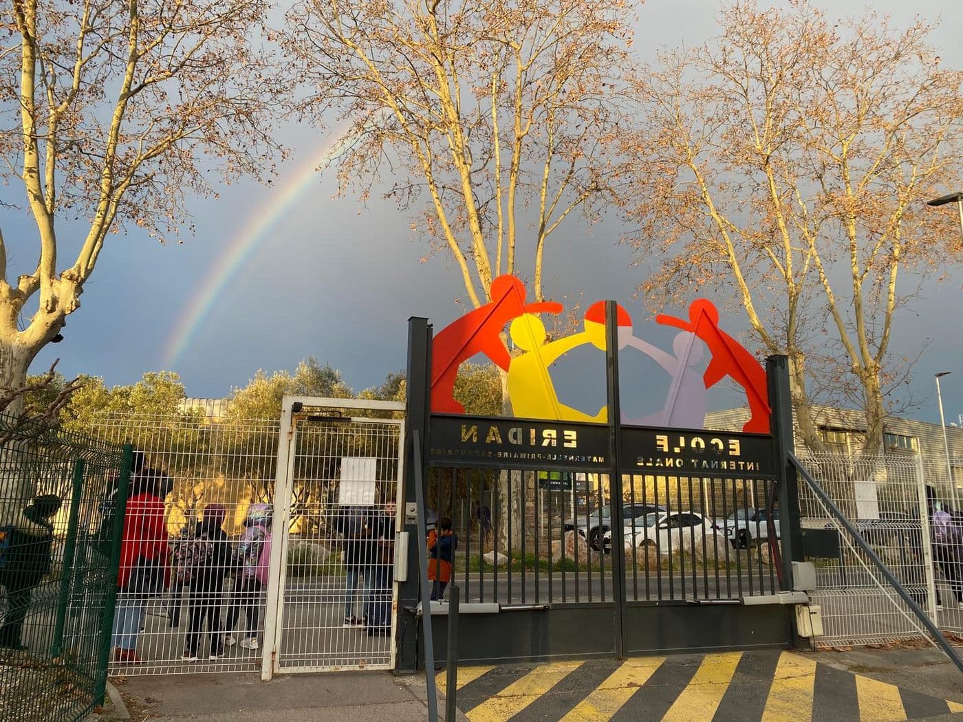 Le portail de l'école vu depuis le campus, sous un arc-en-ciel et des platanes dorés.