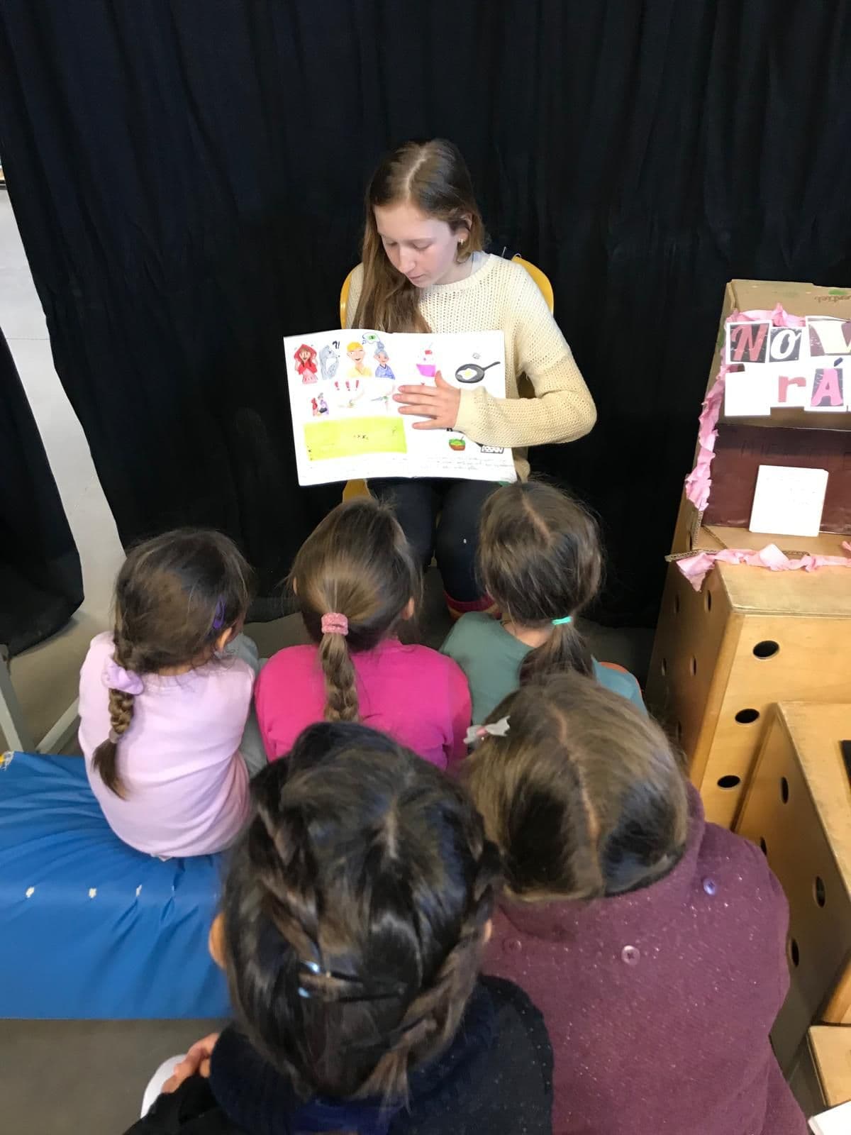 Une grande élève lit un album aux plus jeunes, assis devant elle.