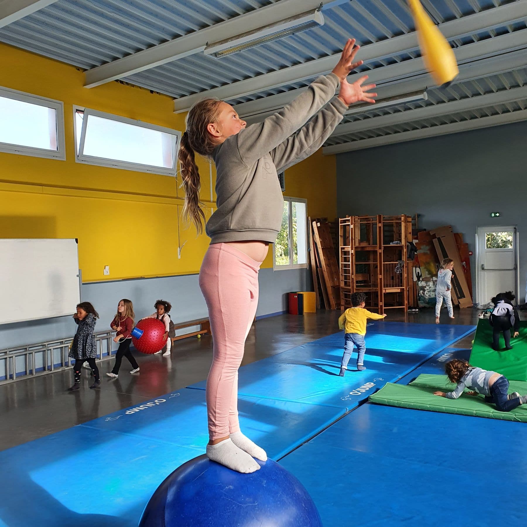 Circus workshop: a pupil balancing on a big ball throws a juggling club.