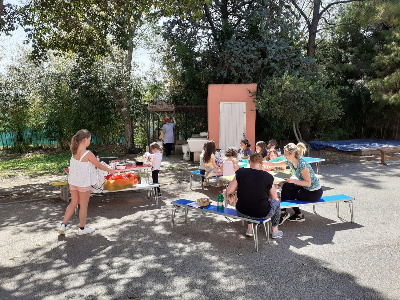 Children and adults sitting at tables in the shade of the trees in the playground.