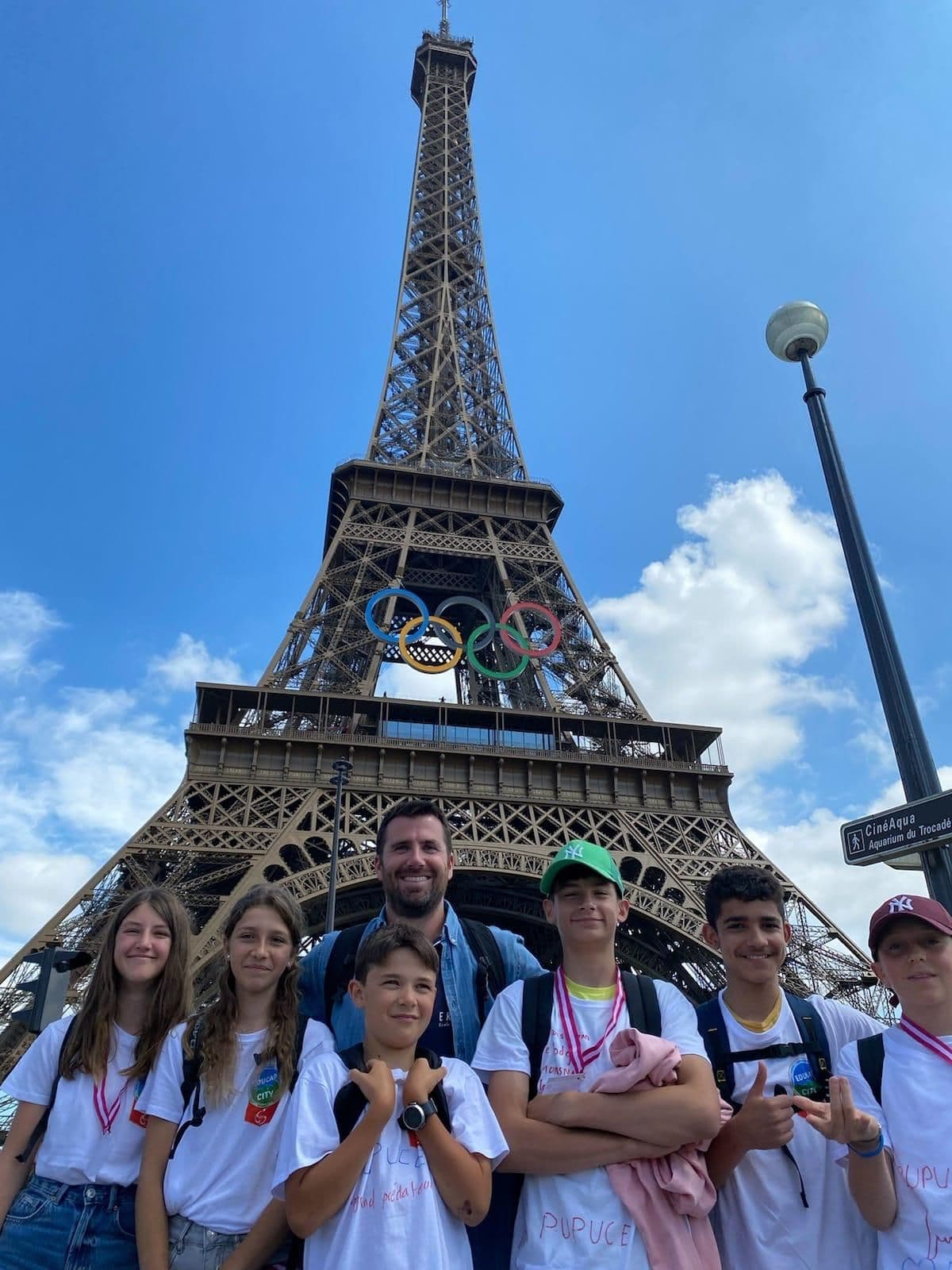 Middle school pupils and their teacher at the foot of the Eiffel Tower, decked with the Olympic rings.